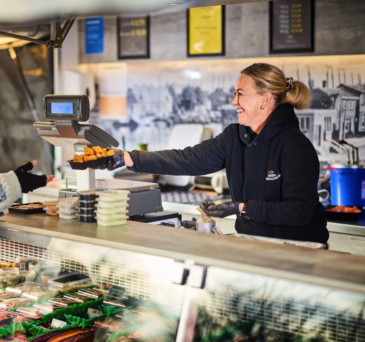 Medewerkster van Gerie's overhandigt met een glimlach een bakje verse kibbeling aan een klant bij de verkoopwagen